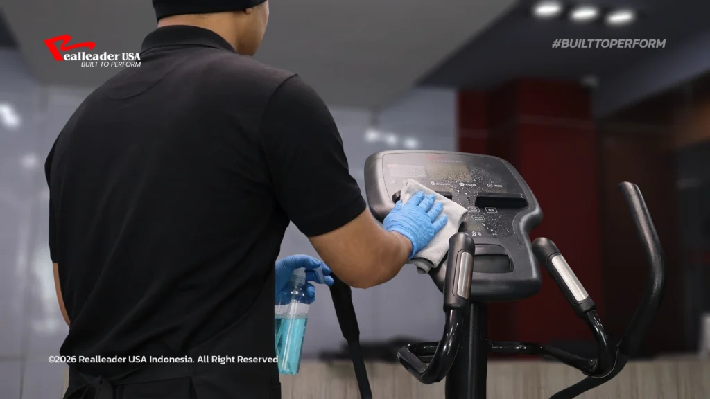 Foto close-up berkualitas tinggi yang memperlihatkan seorang staf gym profesional sedang membersihkan Mesin Cardio Realleader dengan Tips Rawat Alat Gym